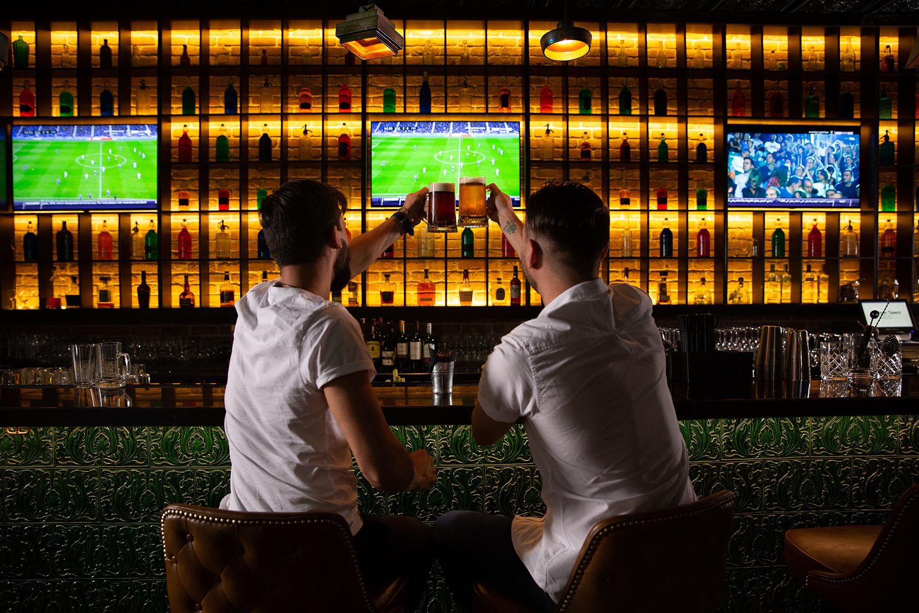 Hombres brindando en un bar con un partido de fútbol de fondo